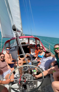 A group of people sitting on a sailboat, smiling and holding drinks, with the ocean and clear sky in the background.