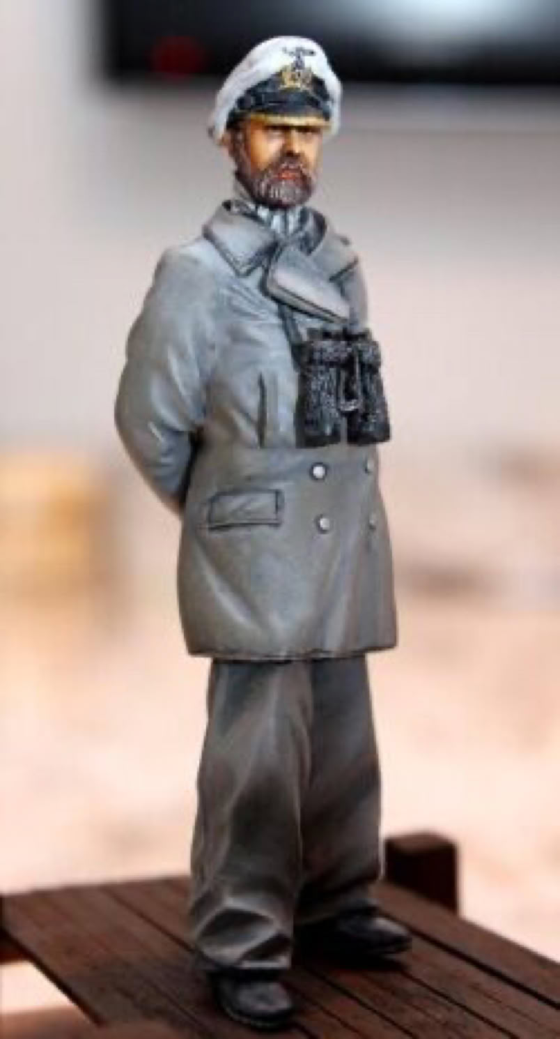 A small figurine of a person in a gray military uniform with a cap and binoculars around the neck, standing on a wooden surface.