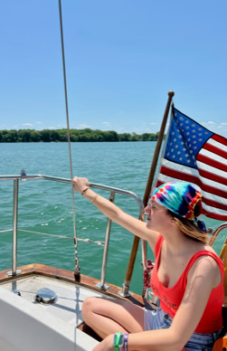 Person wearing a colorful headscarf and red tank top sitting on a boat, with an American flag in the background and water and trees visible.