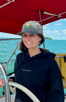 Person wearing a cap and hoodie steering a sailboat with a red canopy, surrounded by blue water and sky.