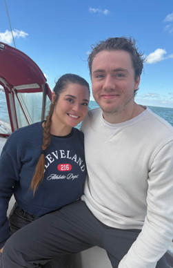 A man and woman sitting together on a boat with a blue sky and ocean in the background. The woman is wearing a navy sweatshirt, and the man is wearing a white long-sleeve shirt.